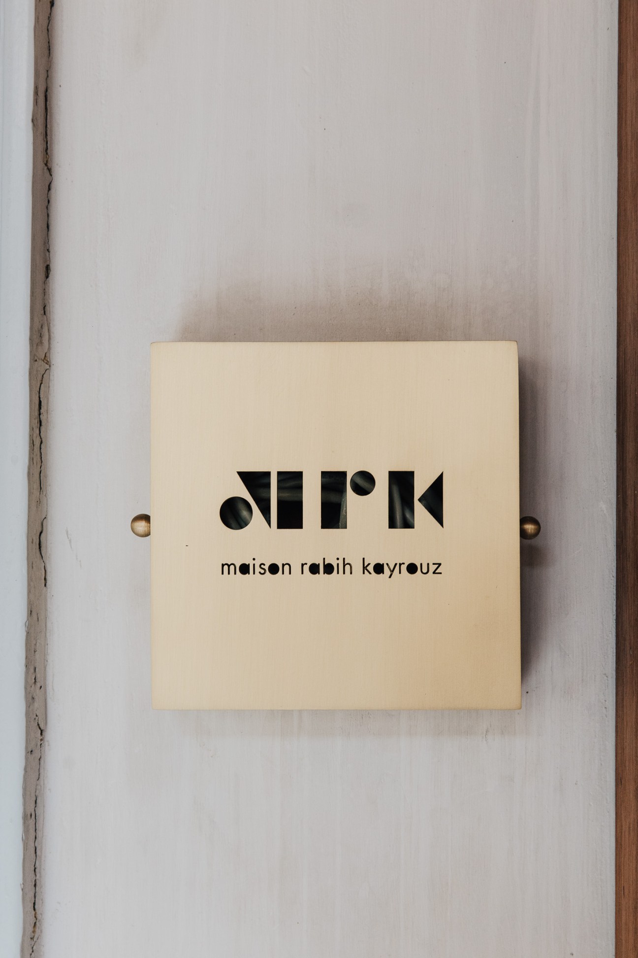Cream-coloured wooden plaque with geometric black logo and text reading 'maison rabih kayrouz' mounted on white wall.