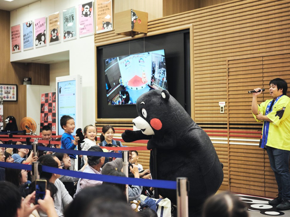 Kumamon performing energetically on a small stage, arms raised as he entertains a crowd of seated fans