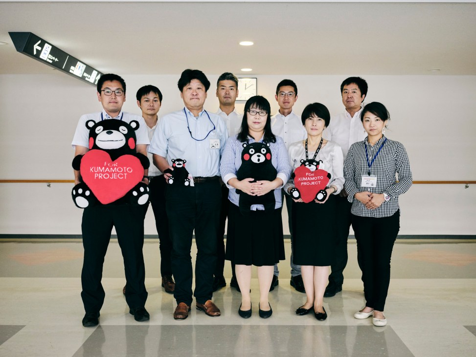Nine Kumamon Group team members in business attire posing together, three holding small Kumamon bear mascot dolls