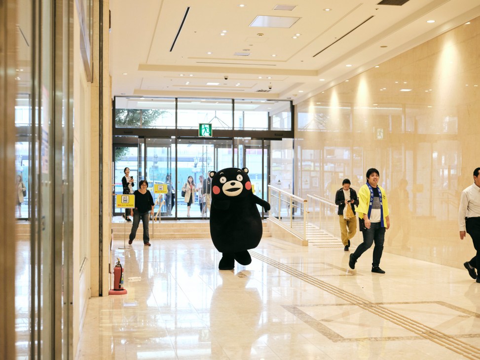 Kumamon running down a modern building hallway with polished floors, captured mid-stride as people watch