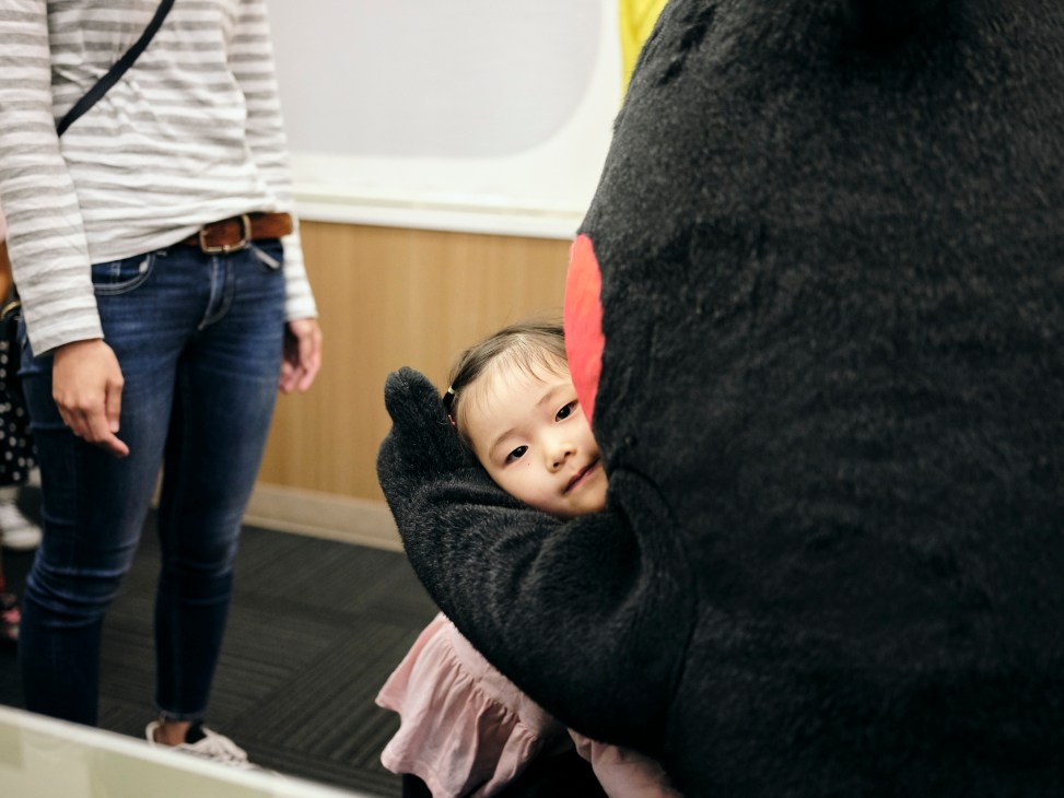 Kumamon embracing a young child in a warm hug while other people stand nearby in what appears to be an indoor setting