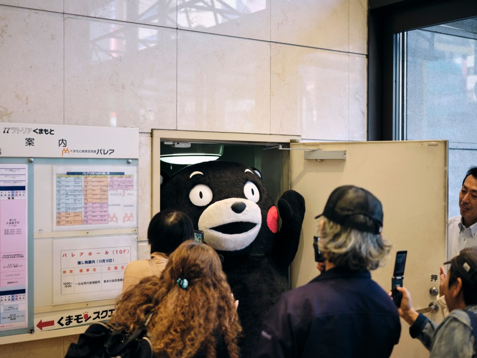 Kumamon mascot emerging from doorway with people gathered around taking photos in what appears to be a service area