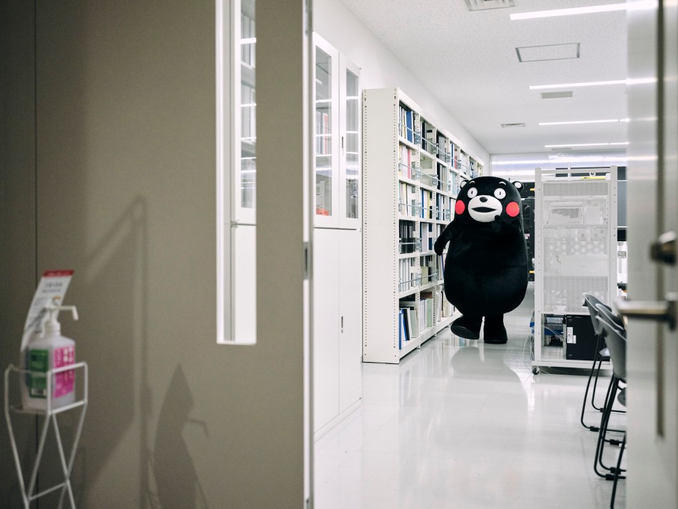 Kumamon mascot walking down a bright library hallway between tall white bookshelves