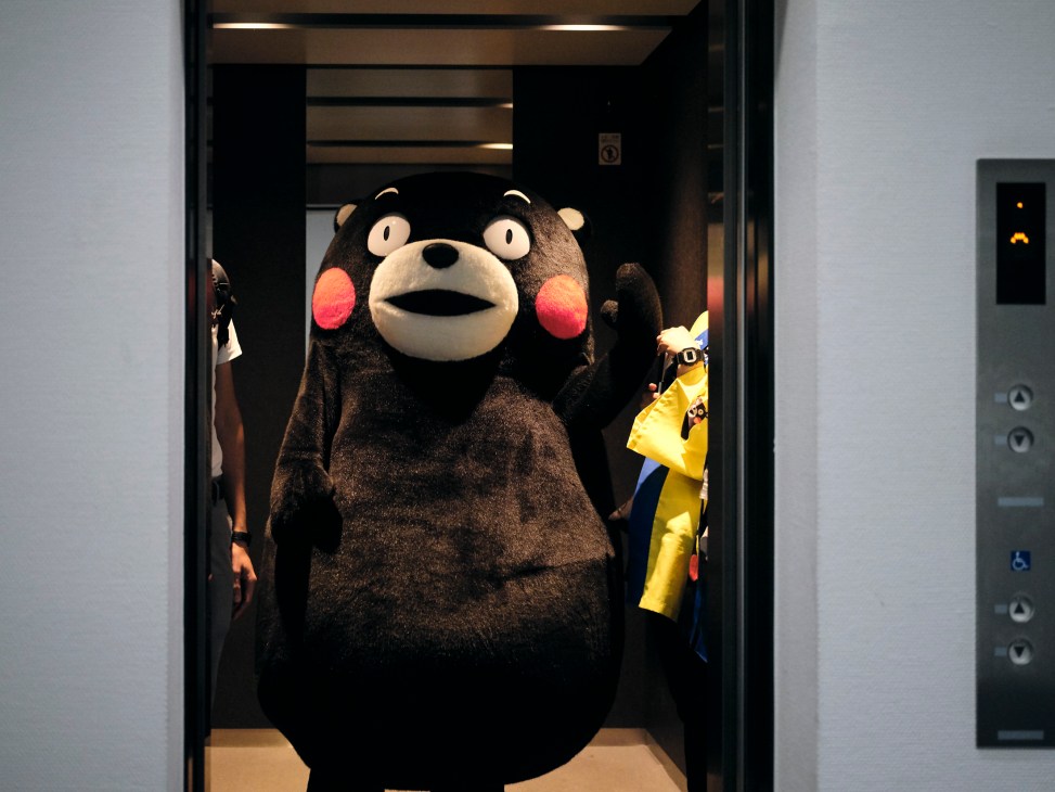 Kumamon mascot character standing in an elevator with other people visible in the background, including someone in a yellow jacket on the right side