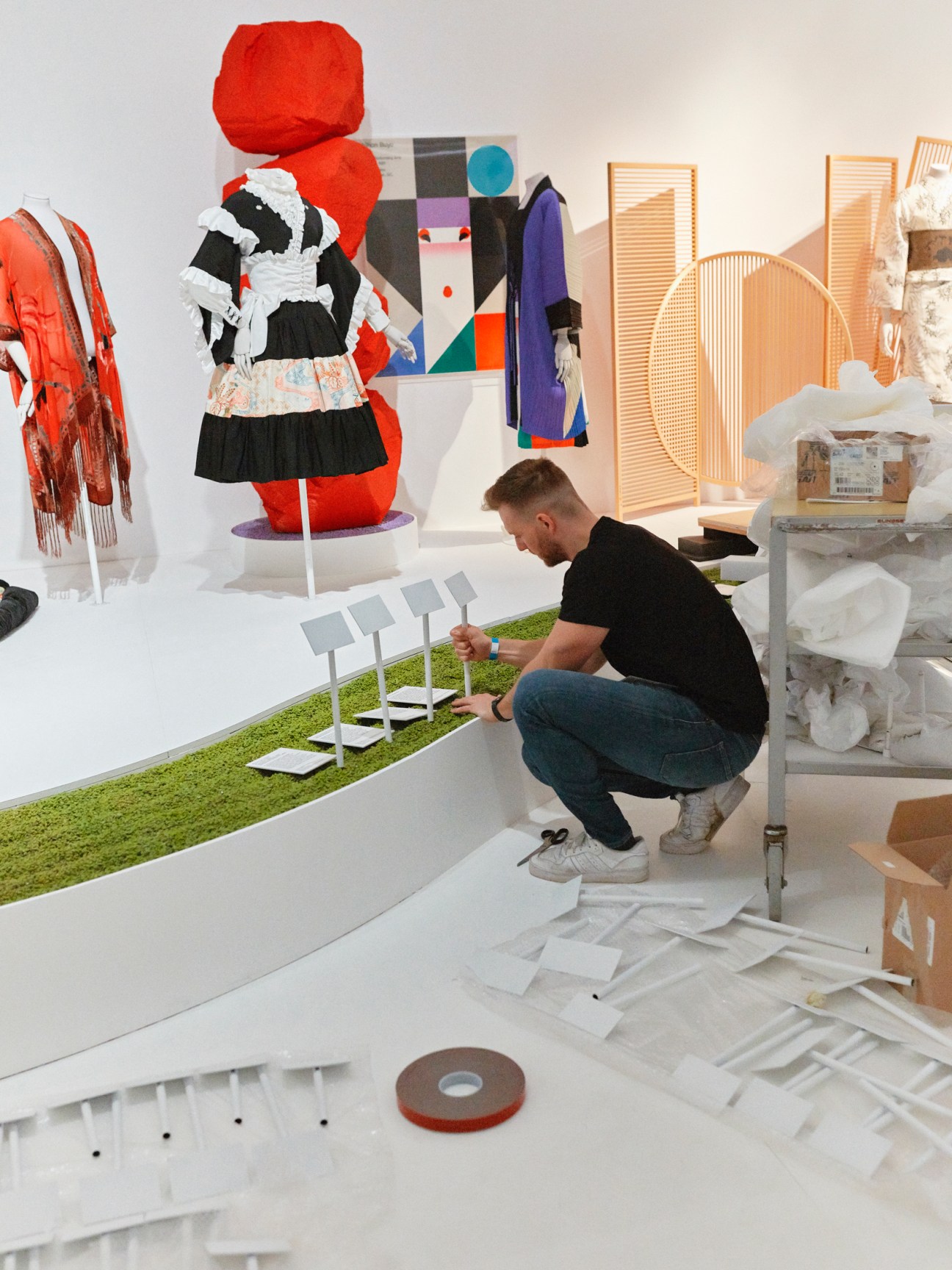 Museum technician kneeling while installing white display stands on artificial grass in exhibition space