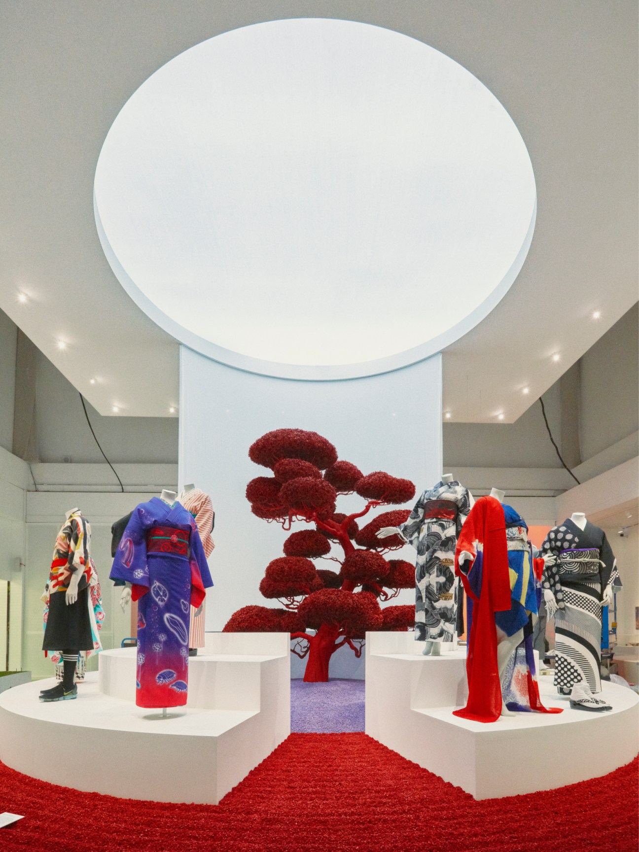 Exhibition display with colorful kimono garments on white platforms around a central red sculptural tree