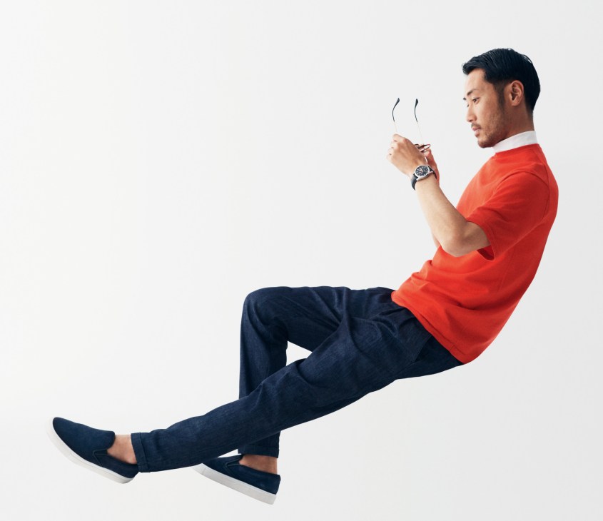 Man in red t-shirt and dark jeans wearing Tag Heuer watch and Ray-Ban glasses, holding pen against white background.