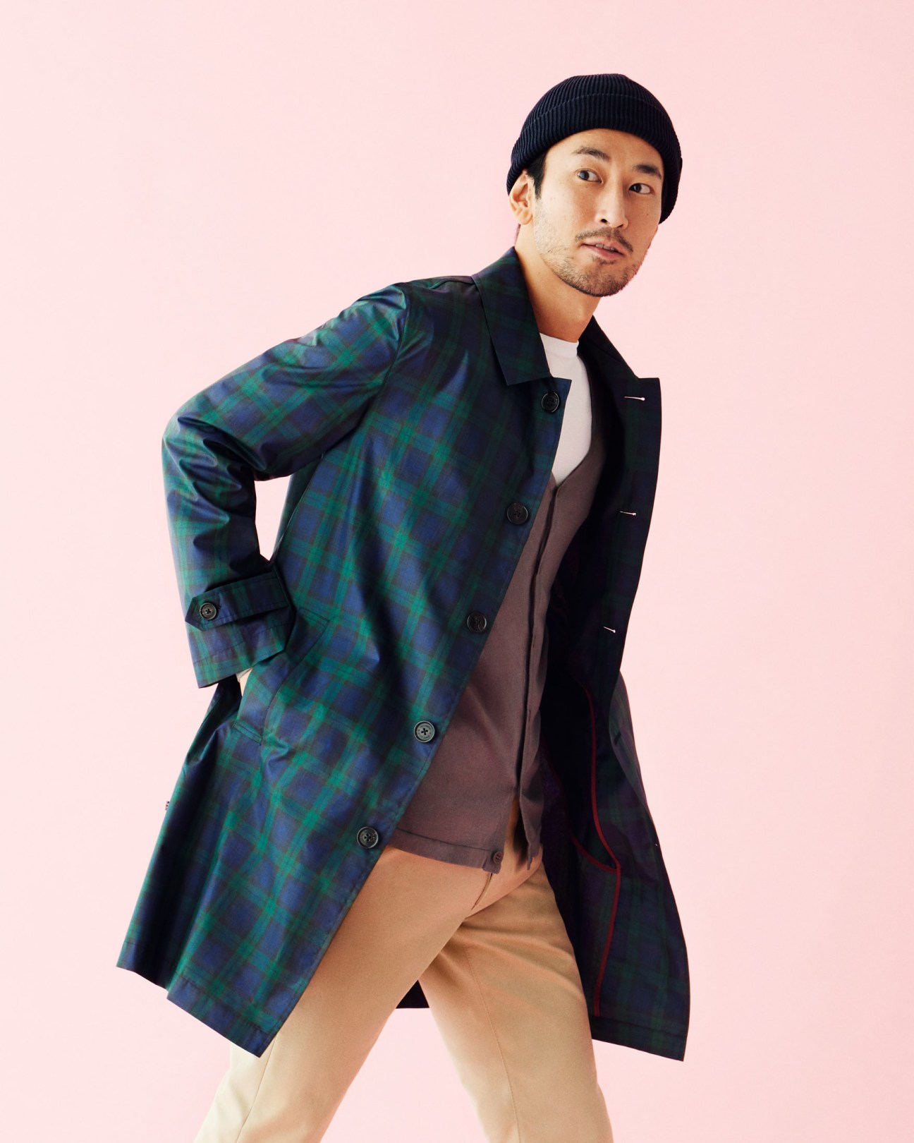 Man wearing plaid coat by Hackett London, navy beanie, and tan trousers against pink background.