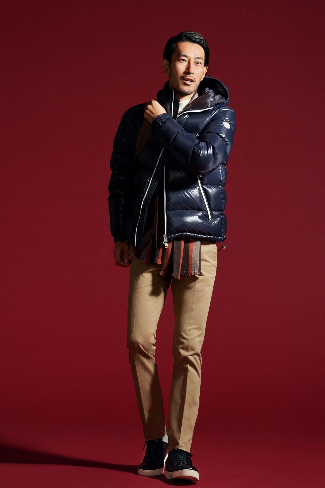 Man in black puffer jacket with grey turtleneck and beige trousers wearing striped scarf against red background