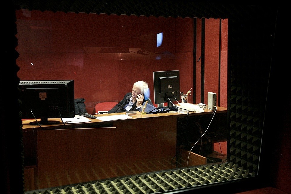 An older man sits at a desk in a red-walled recording studio, speaking on a telephone.