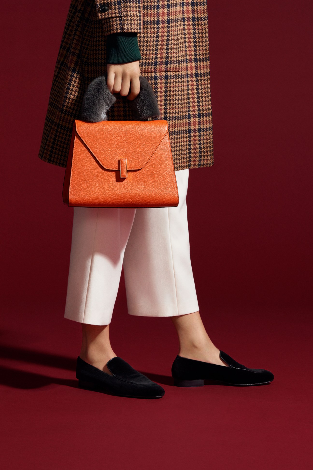 Woman in camel plaid coat holding orange Valextra bag with gray fur handle detail over white trousers and black loafers.
