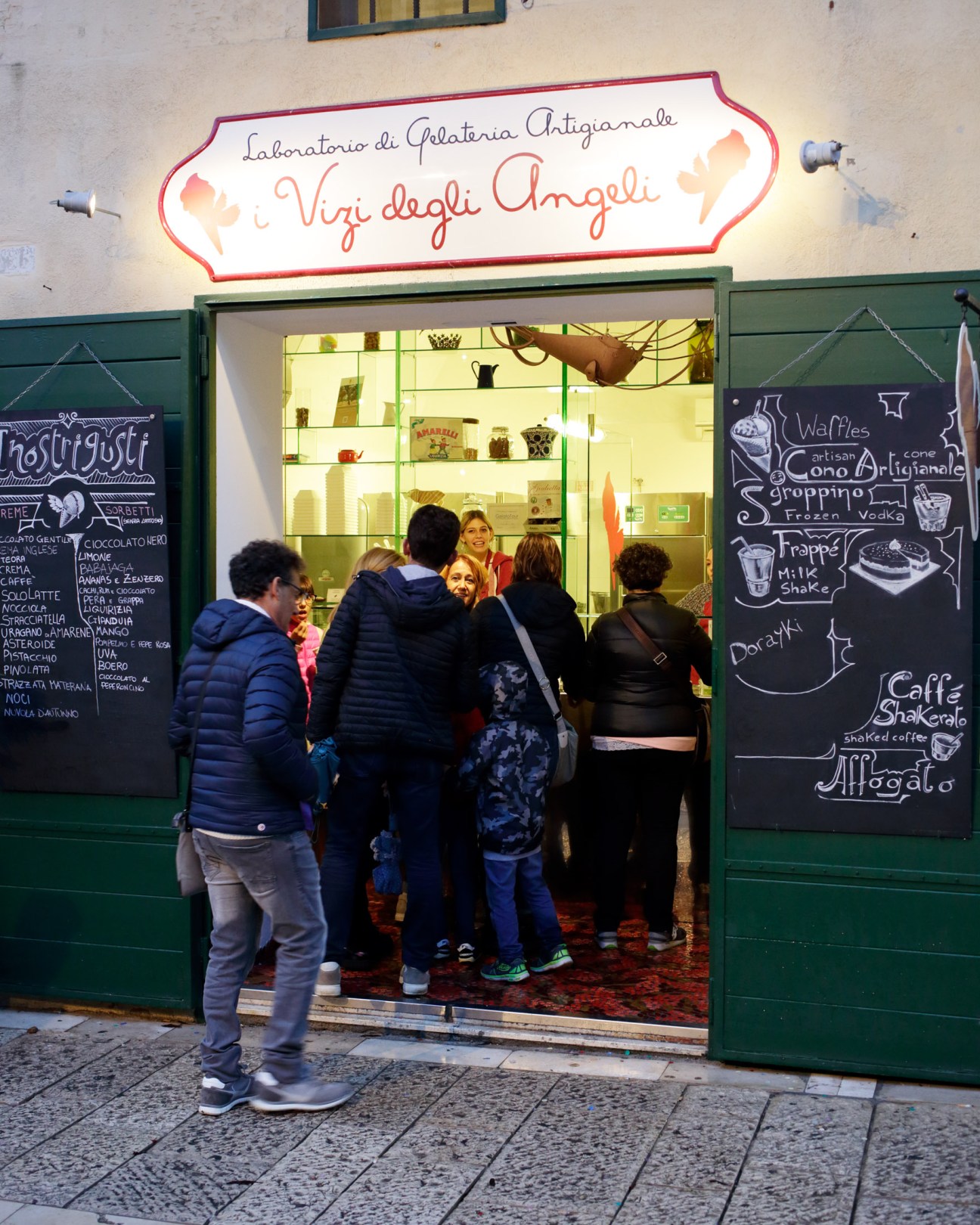 Storefront of Vizi degli Angeli, a gelato shop in Matera with neon signage and customers at the entrance.