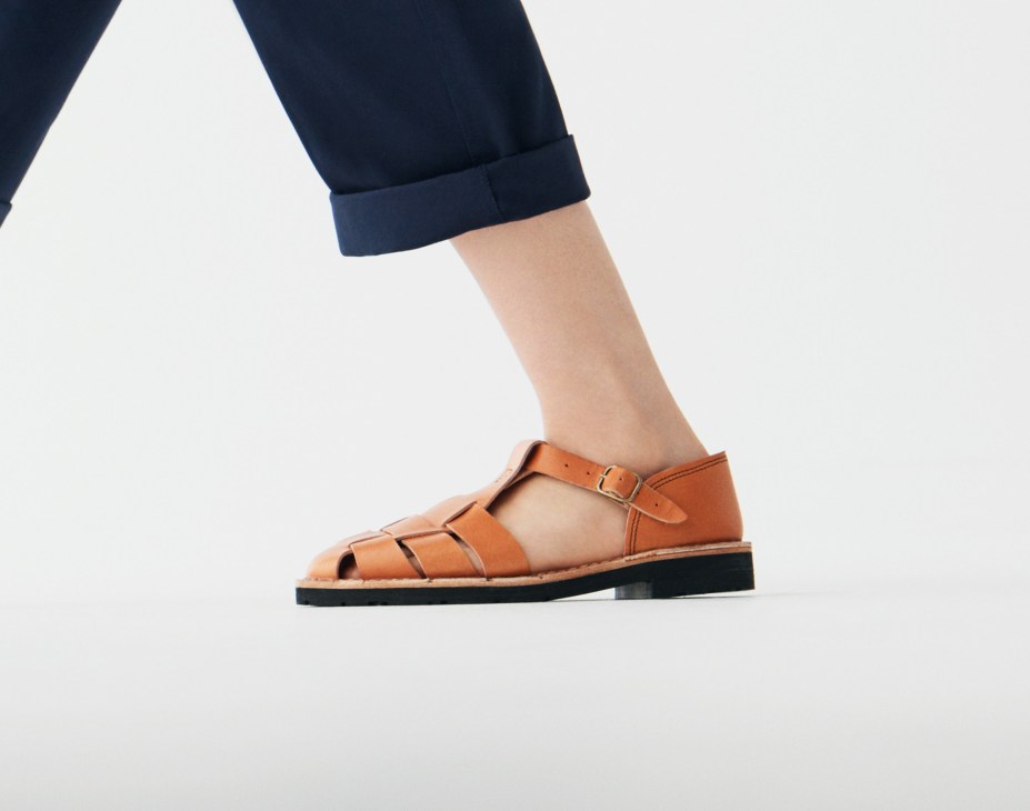 Person wearing navy trousers and tan leather sandals with black sole against white background.
