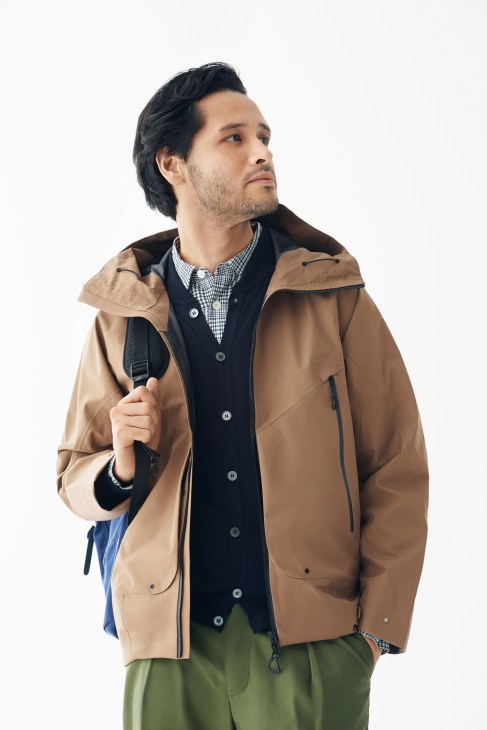 Man wearing a tan Goldwin jacket, navy Zanone cardigan, and checkered APC shirt with green Still By Hand trousers, holding a Kiruna backpack.