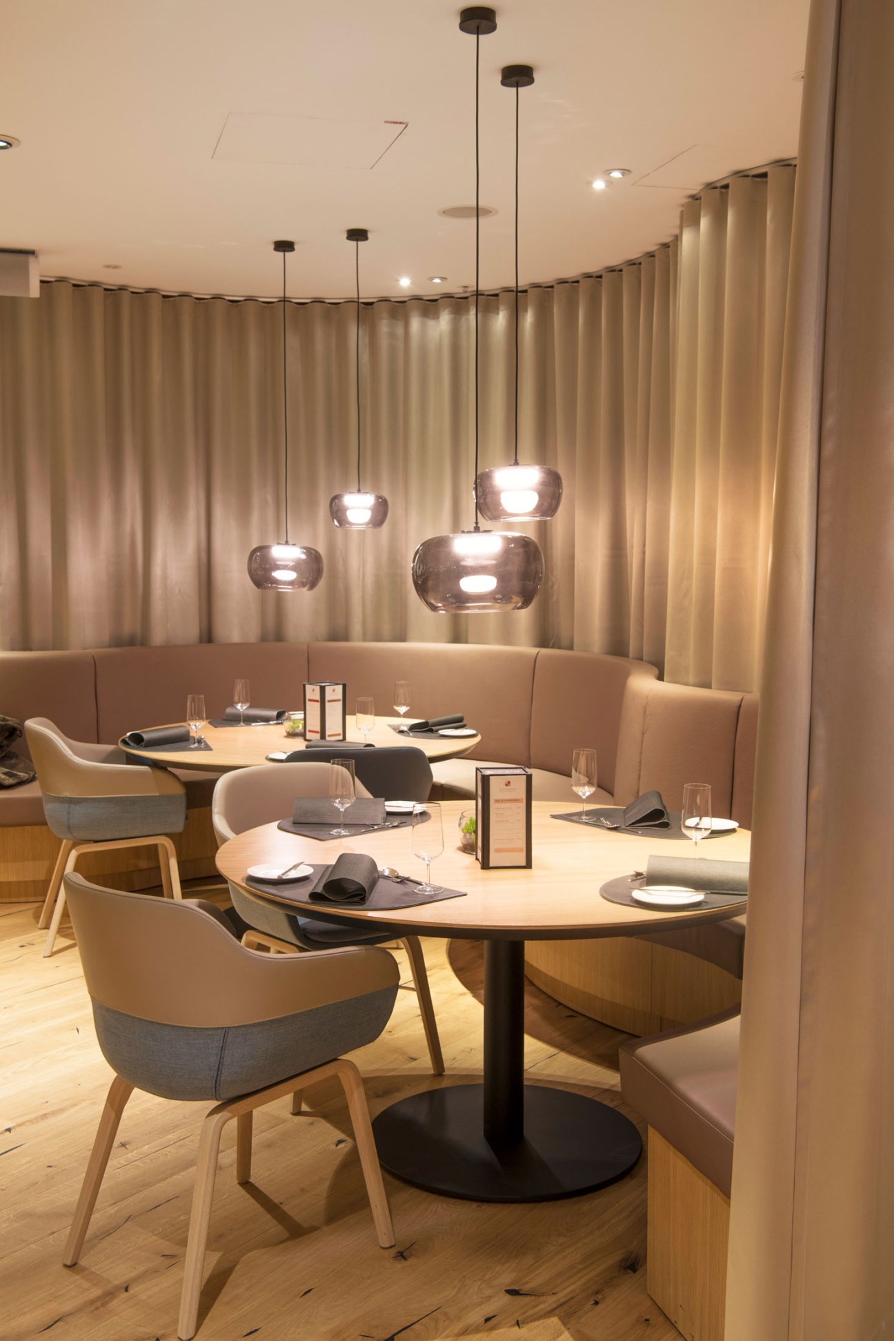 Modern restaurant interior with round tables, beige upholstered seating, pendant lighting and floor-to-ceiling curtains.