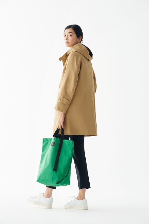 Woman wearing a beige Orchival coat, black Arket trousers, white Santoni trainers, and holding a green Bagsinprogress bag.