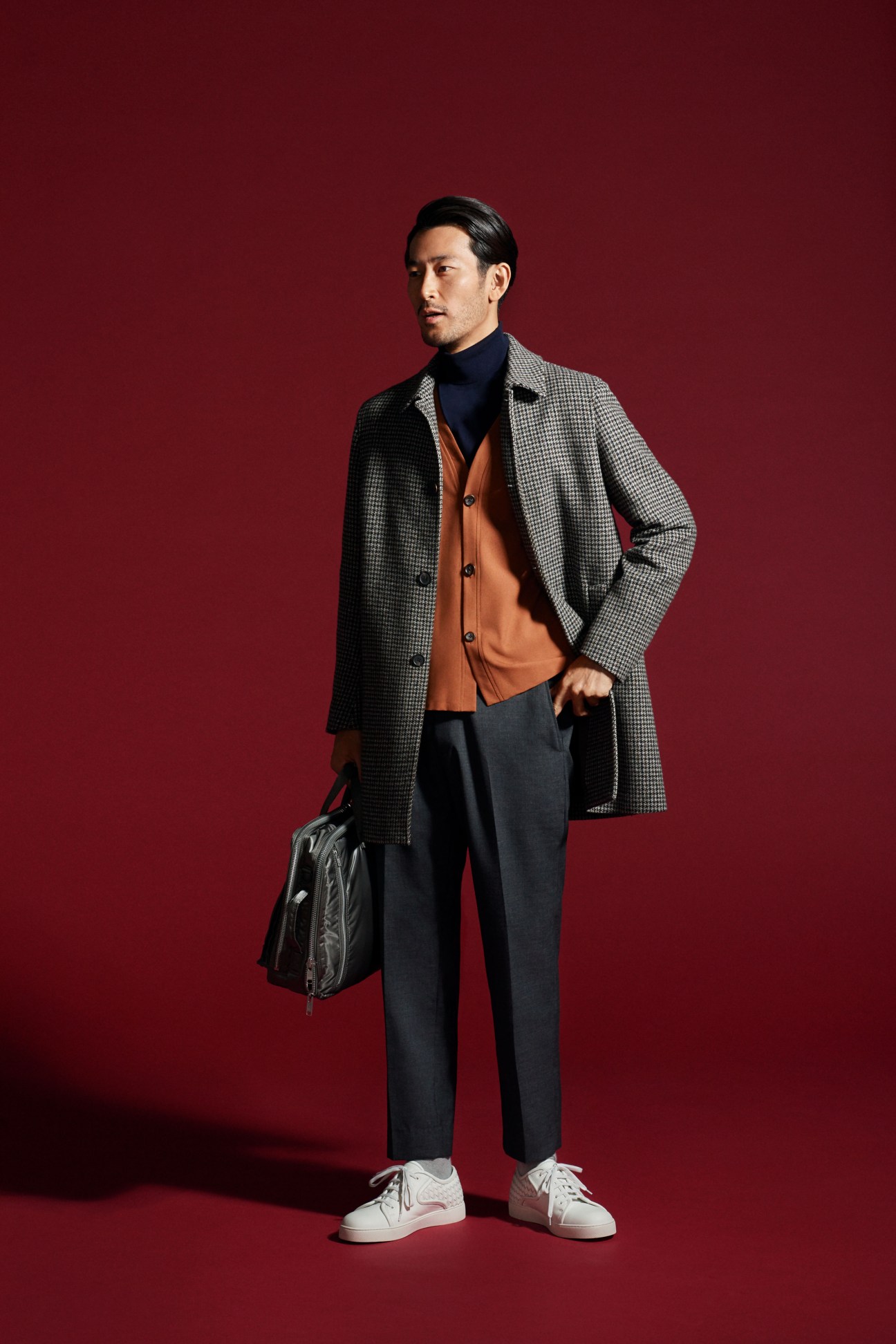 Man in brown coat with grey cardigan over black turtleneck and dark trousers carrying black bag wearing white sneakers against red background