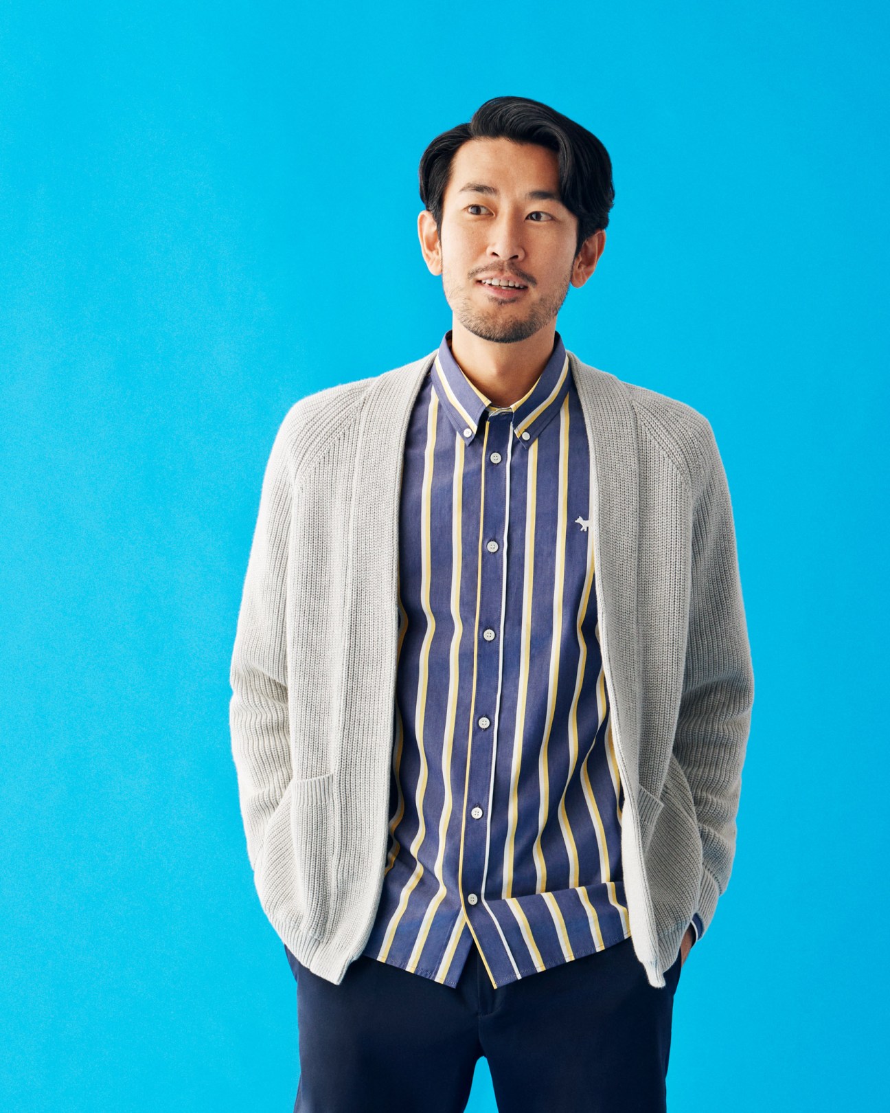 Man wearing a cream cardigan, navy striped shirt, and dark trousers against a blue background.