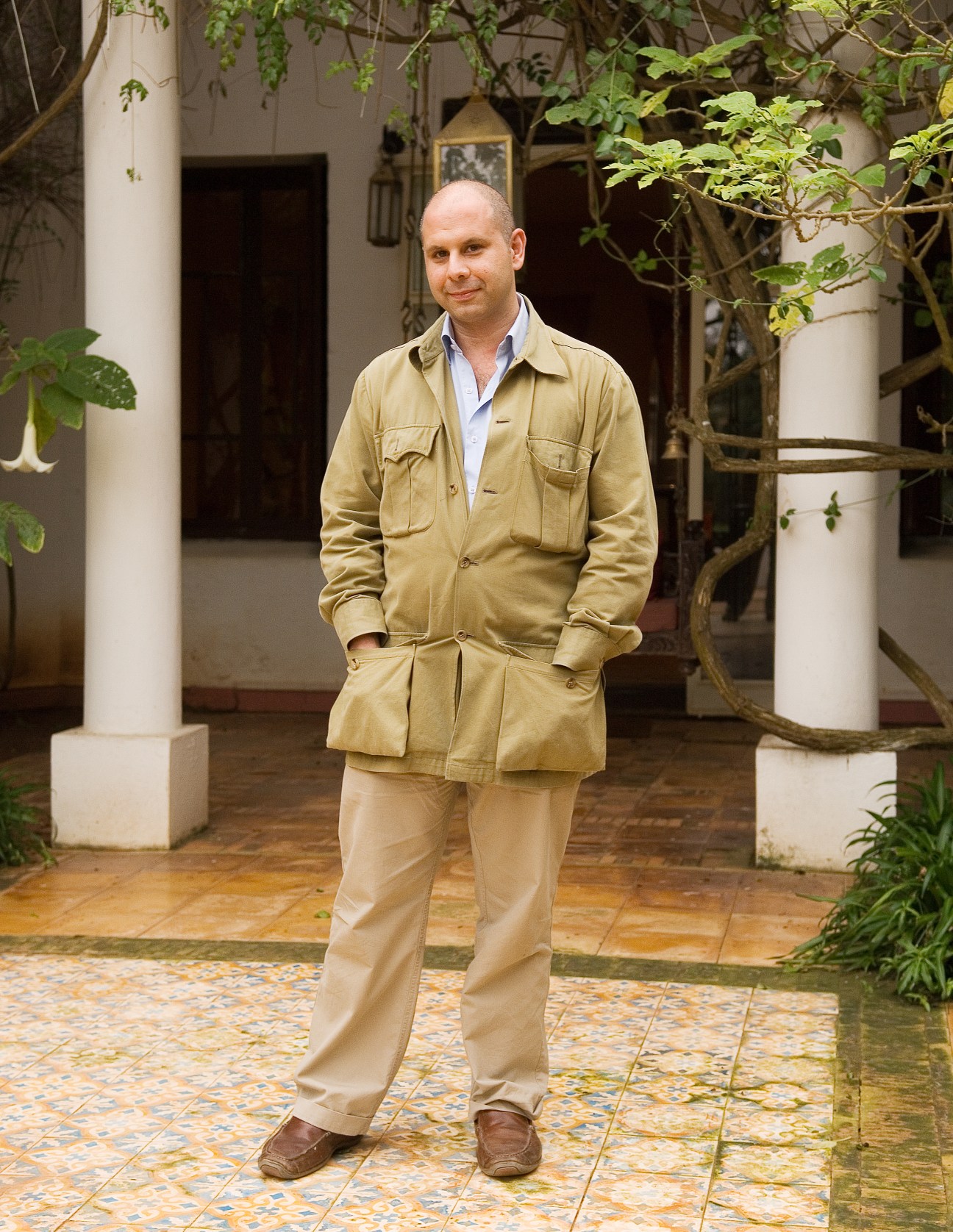 Man in tan jacket standing in a courtyard with white columns and climbing vines.