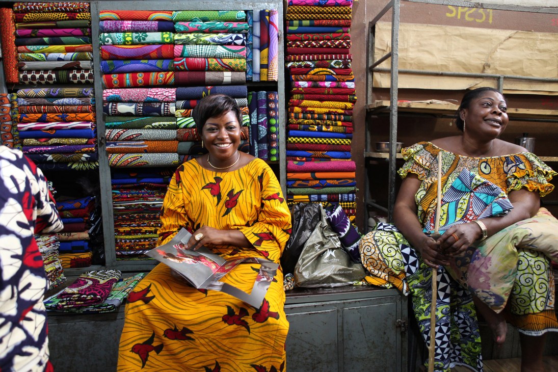 Saleswoman in yellow patterned dress sits with fabric samples displayed behind her at a Vlisco market stall.