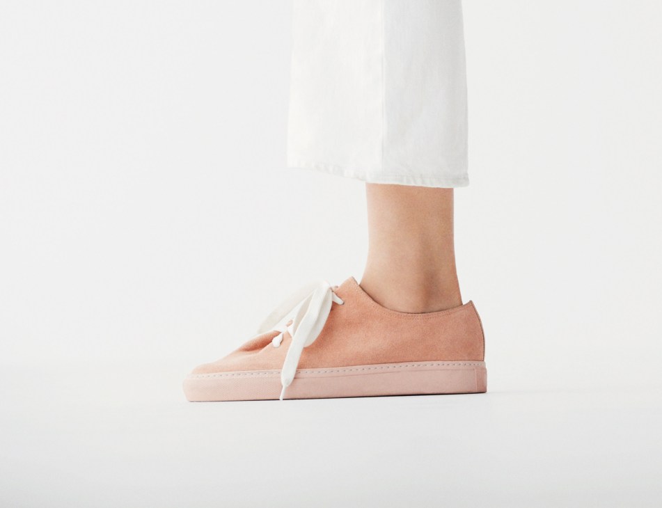 Close-up of feet wearing pale pink trainers with white laces and white cropped jeans against a light background.