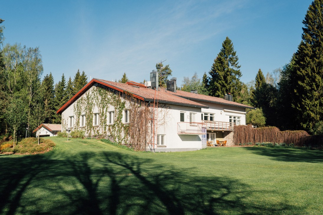 White modernist country house with red-trimmed roof surrounded by forest and manicured lawn.