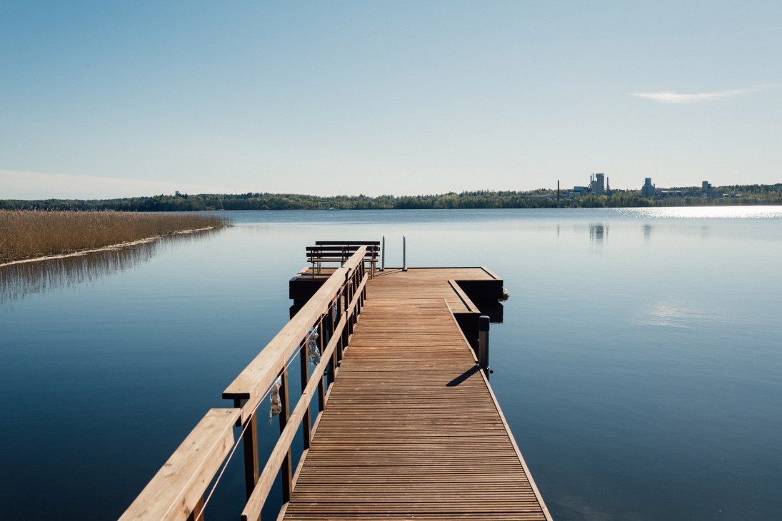 Wooden jetty extending into calm blue lake with forest and city skyline in distance.