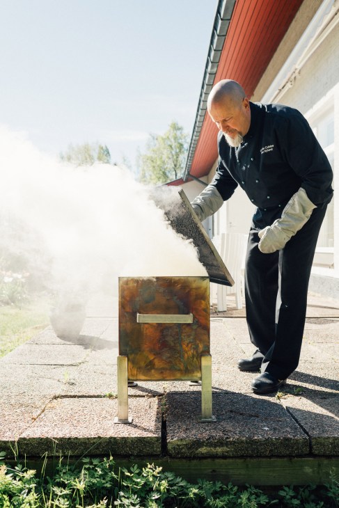 Man in dark jacket and work gloves tends to a traditional Finnish smoke sauna by a lake.