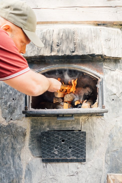 Person in red shirt and beige cap stoking a fire in a traditional Finnish smoke sauna.