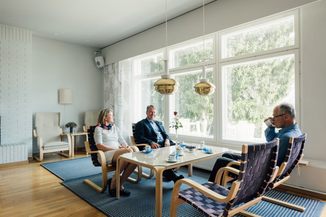 Three people seated in a bright modern meeting room with large windows overlooking forest, gold pendant lights, and wooden furniture.