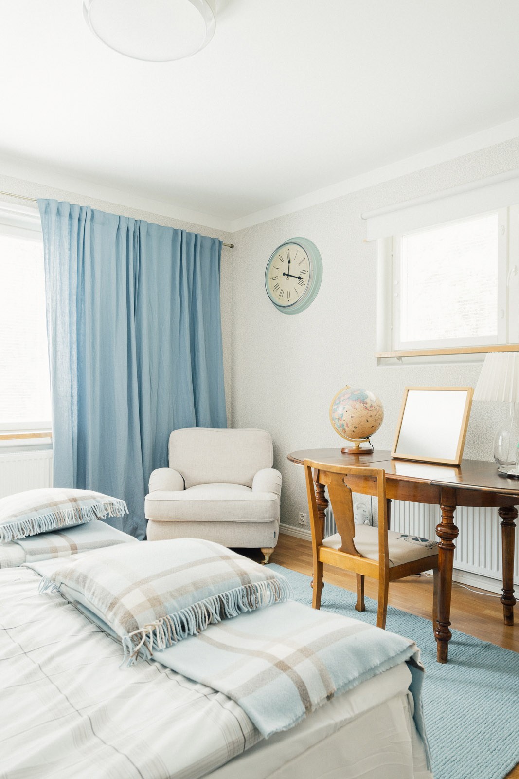 A bright, minimalist bedroom with a cream armchair, wooden desk, blue curtains, and striped bedding in soft tones.