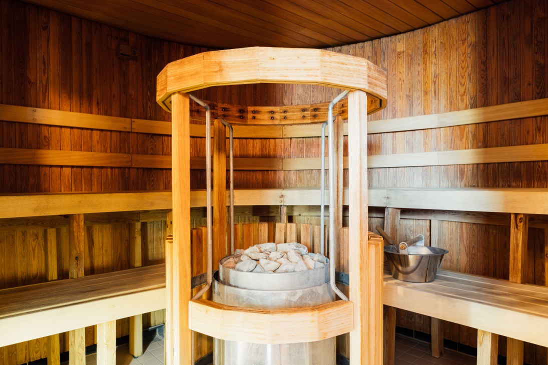 Interior of a wooden smoke sauna with benches, a central stove filled with stones, and metal bucket for water.