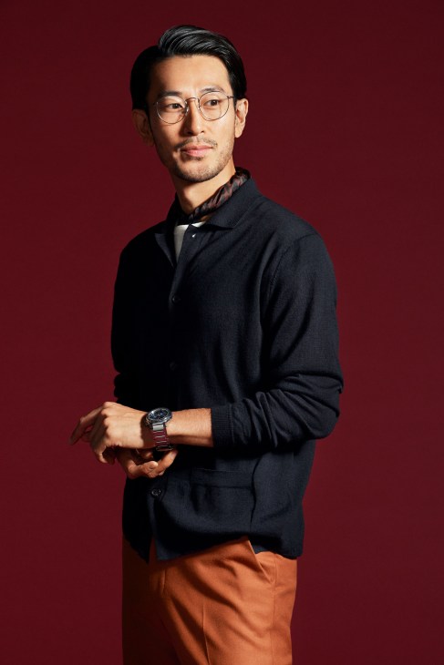 Man in brown cardigan with white t-shirt and dark trousers wearing glasses and scarf with watch standing against white background