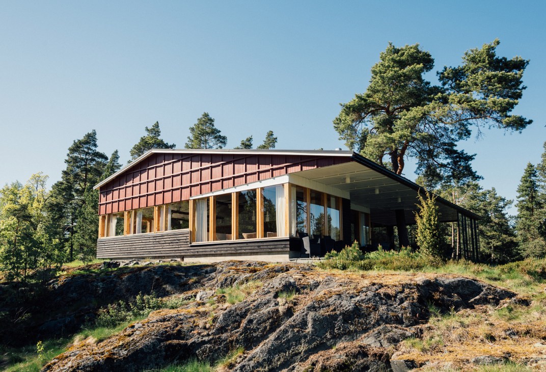 Modern wood-clad pavilion with terracotta roof surrounded by pine trees and rocky terrain in Finnish forest setting.