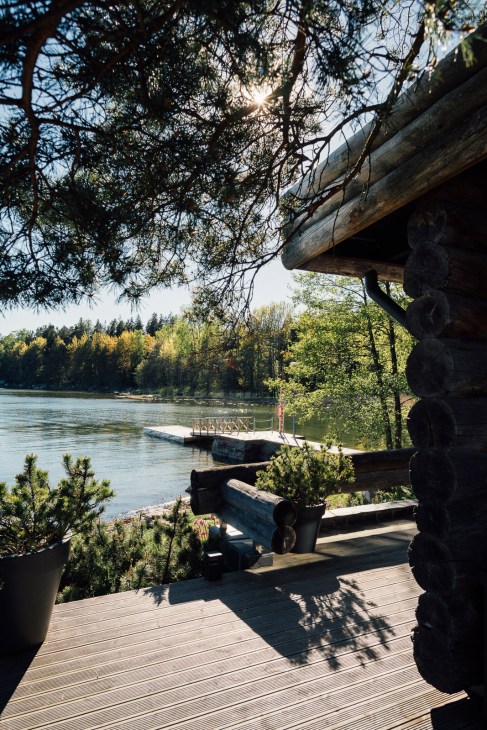 Wooden deck overlooking a lake with logs and sauna structure, forest and jetty visible across the water.