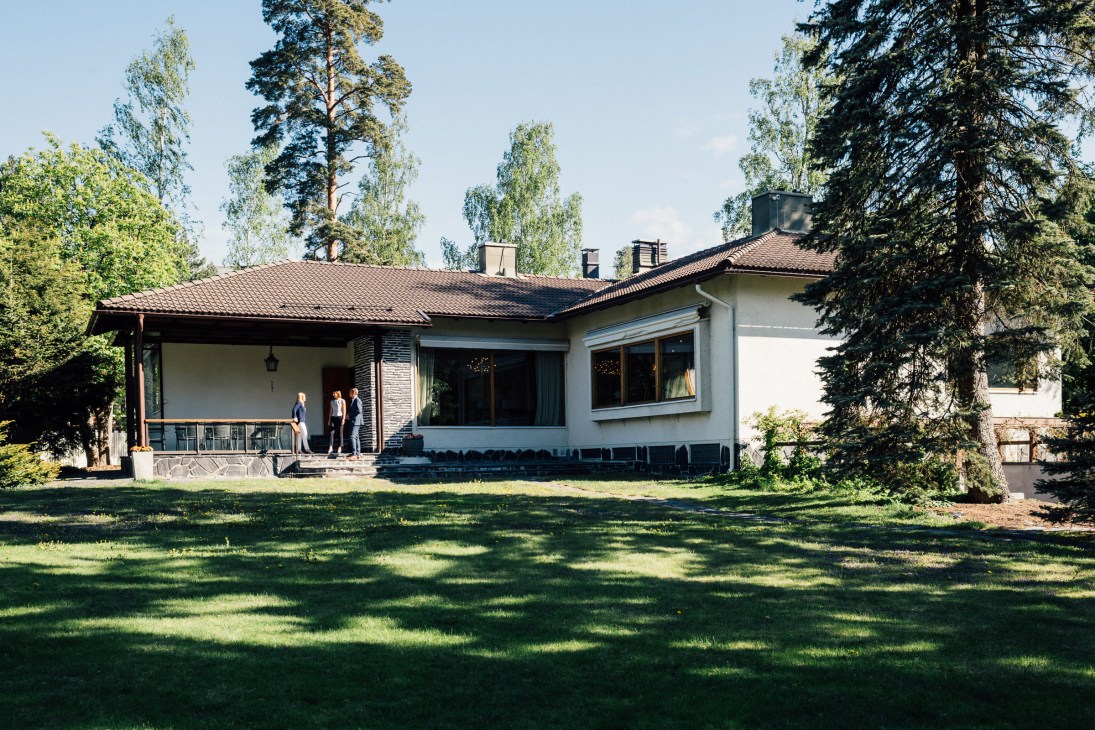 Mid-century modernist country house with white walls and brown roof surrounded by forest and lawn, with people on the entrance porch.