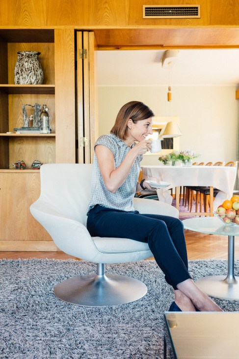 Woman sitting in white modern chair holding a cup in a bright minimalist living room with wooden shelving and warm lighting.