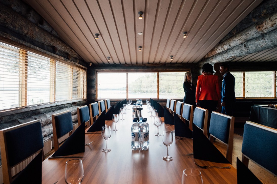 Long dining table set with glasses and water bottles in a modernist wood-paneled room with large windows overlooking water.