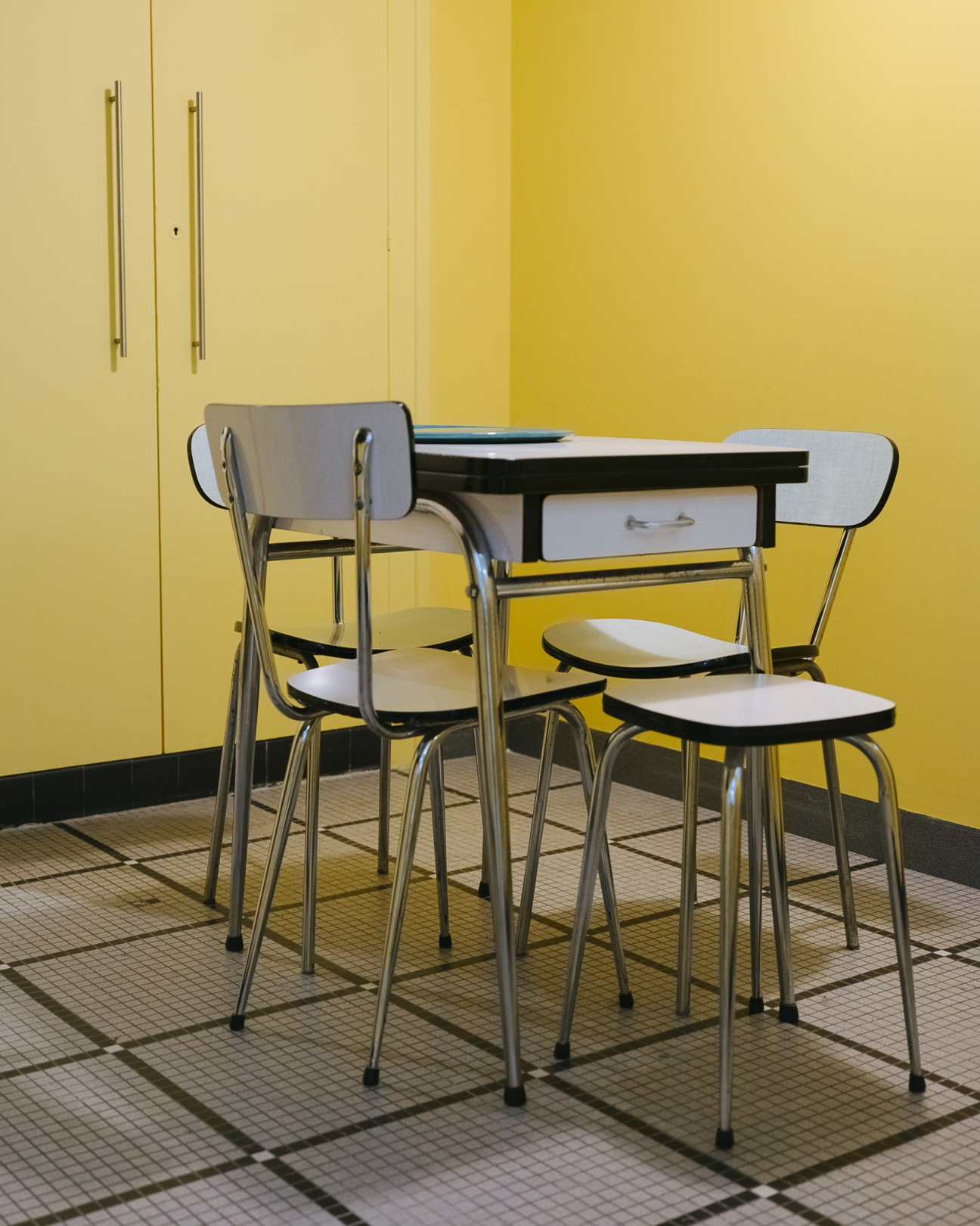 Mid-century Formica chairs and desk with metal legs in a yellow-walled room with checkered flooring.