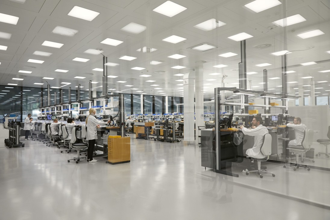Workers in white lab coats assemble watches at individual stations in a bright, modern manufacturing facility with grid ceiling lights.