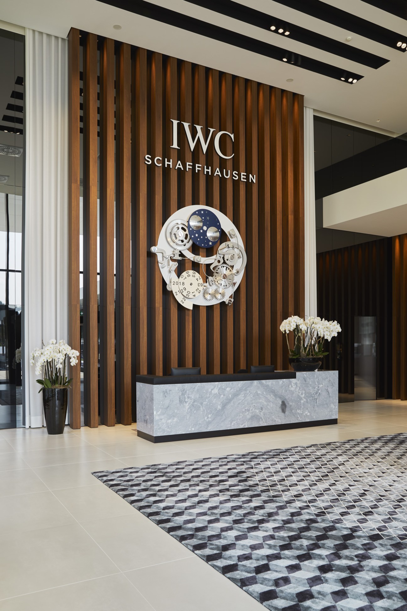 IWC Schaffhausen visitor centre entrance with wooden slat wall, illuminated watch mechanism display, and marble reception desk.