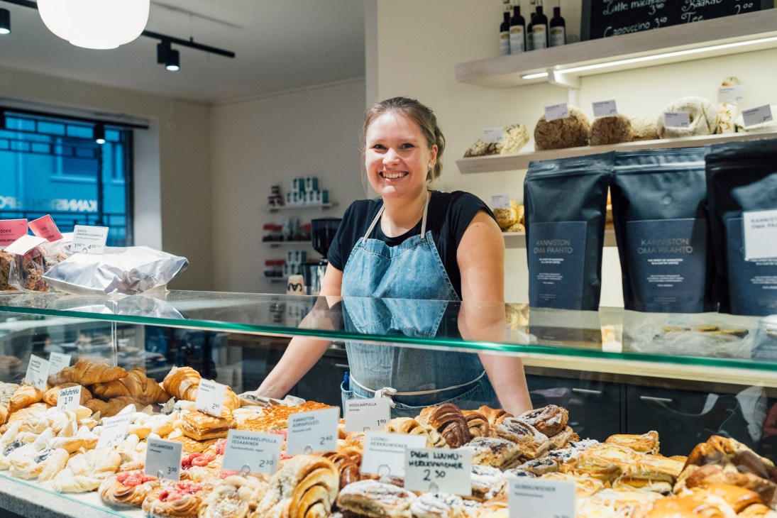 Smiling bakery worker stands behind glass counter display of fresh pastries and buns.