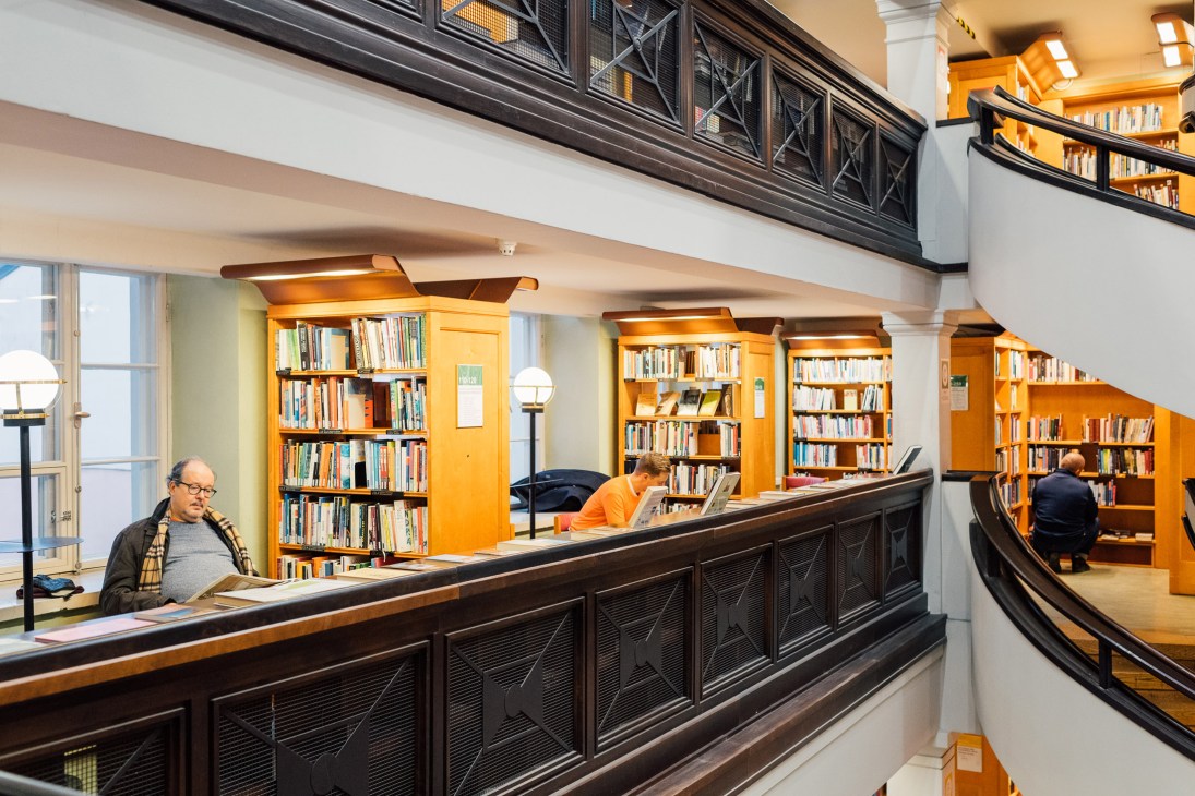 Interior of Rikhardinkatu library in Helsinki with wooden shelving, a dark counter desk, and patrons browsing books.