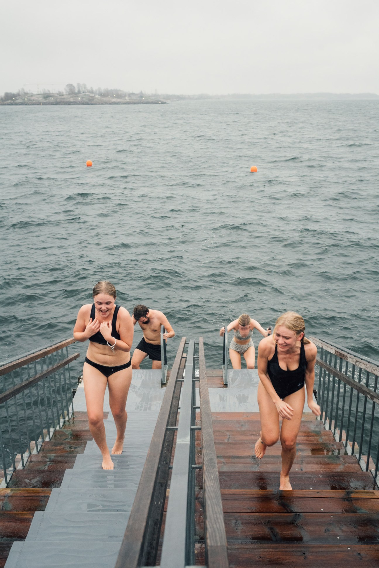 Four people running on a wooden pier into cold water with orange buoys visible in the distance.