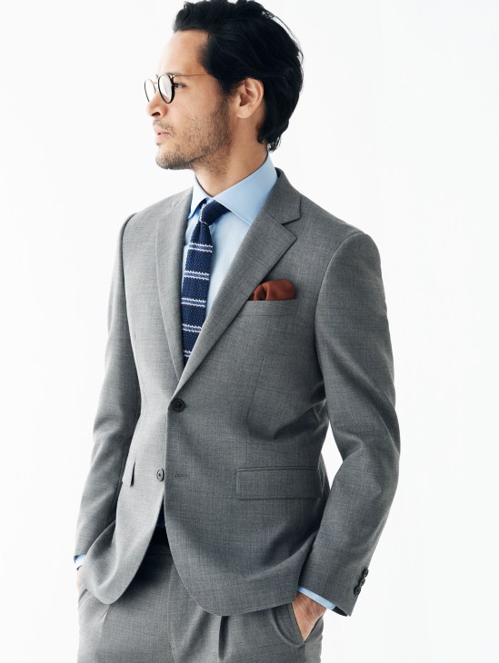Man in gray suit with light blue shirt, patterned tie, and round glasses, styled for business travel.