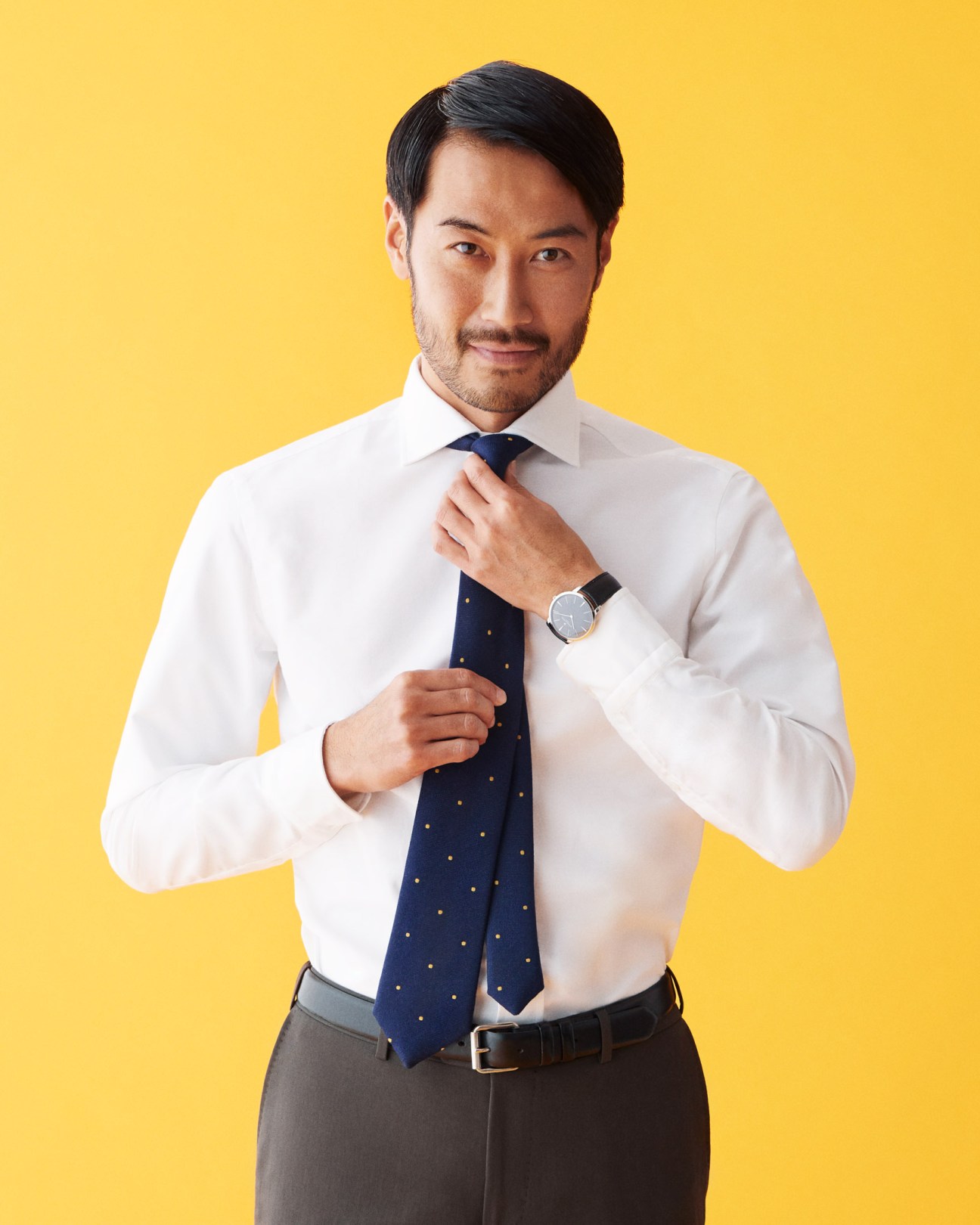Man in white shirt and navy polka dot tie adjusting his tie against yellow background