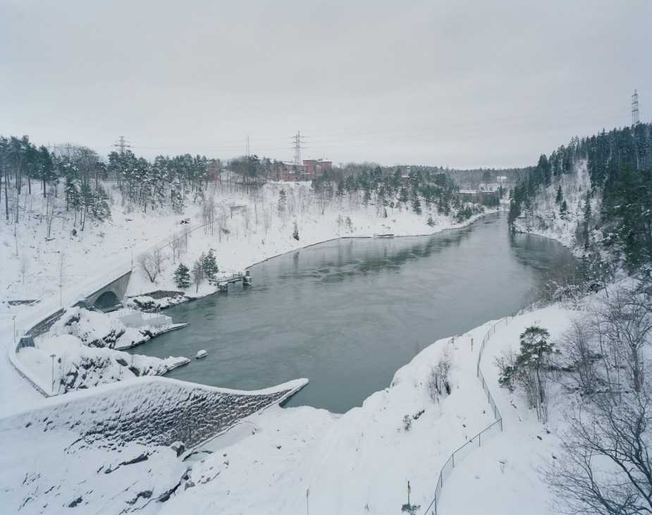 Snow-covered landscape with a river bend, bare trees, industrial buildings, and power lines in winter.