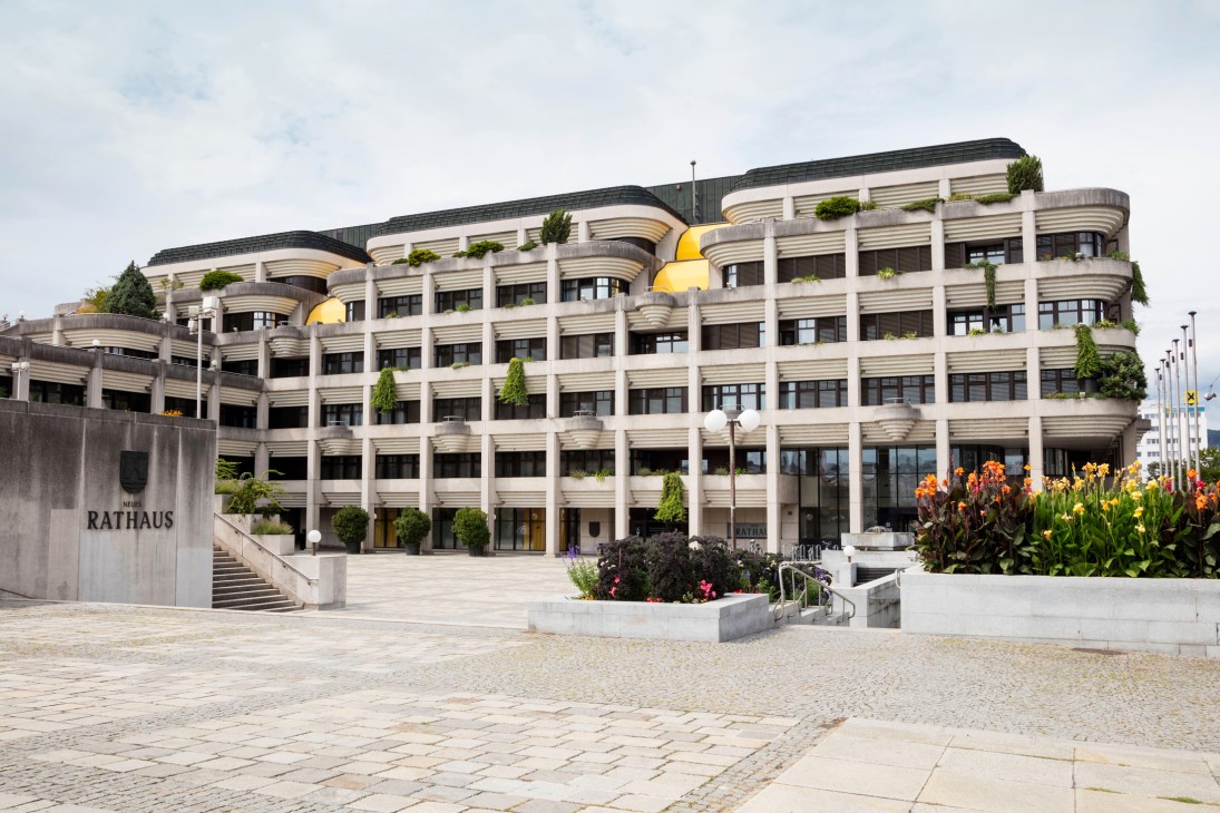 Modern curved concrete building with terraced levels, golden dome features, and landscaped plaza with colorful flowers