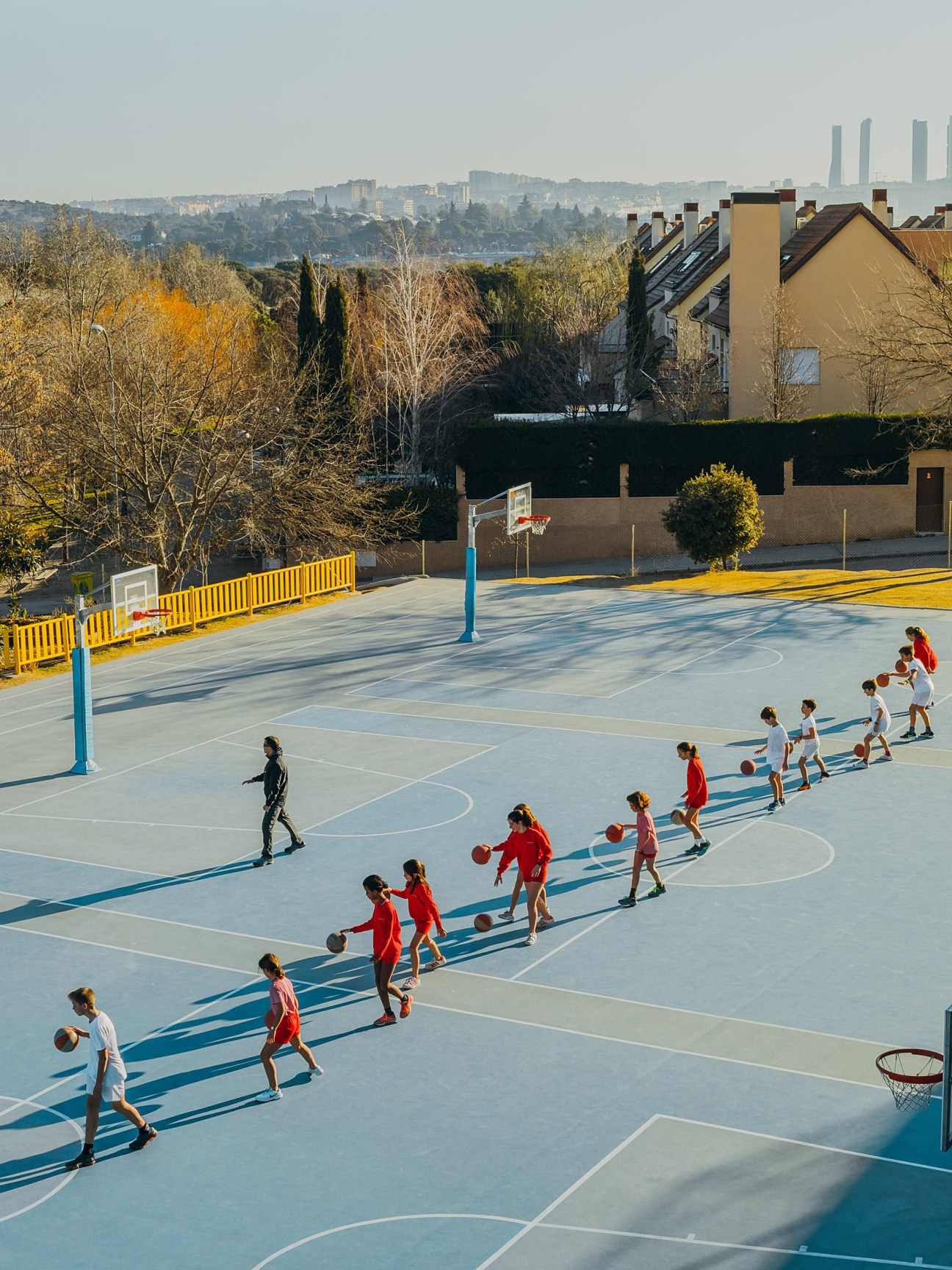 Students in red and white uniforms practicing basketball on an outdoor court with Madrid's skyline visible in the distance.