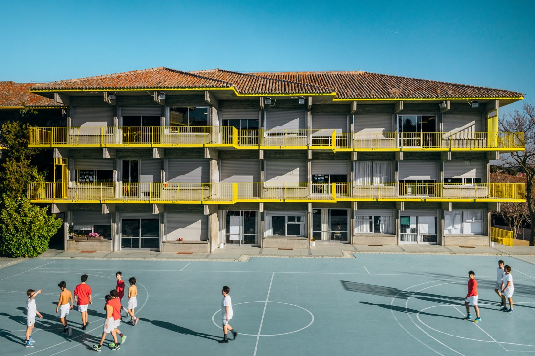 Children playing basketball on an outdoor court in front of a three-story brutalist building with yellow railings and terracotta roof tiles.
